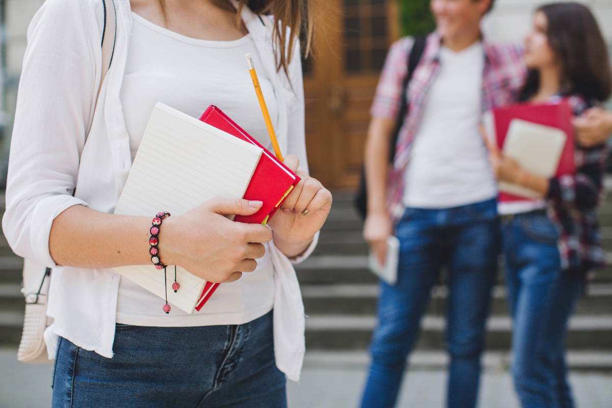 A woman holding notebooks and standing in front of a college building.